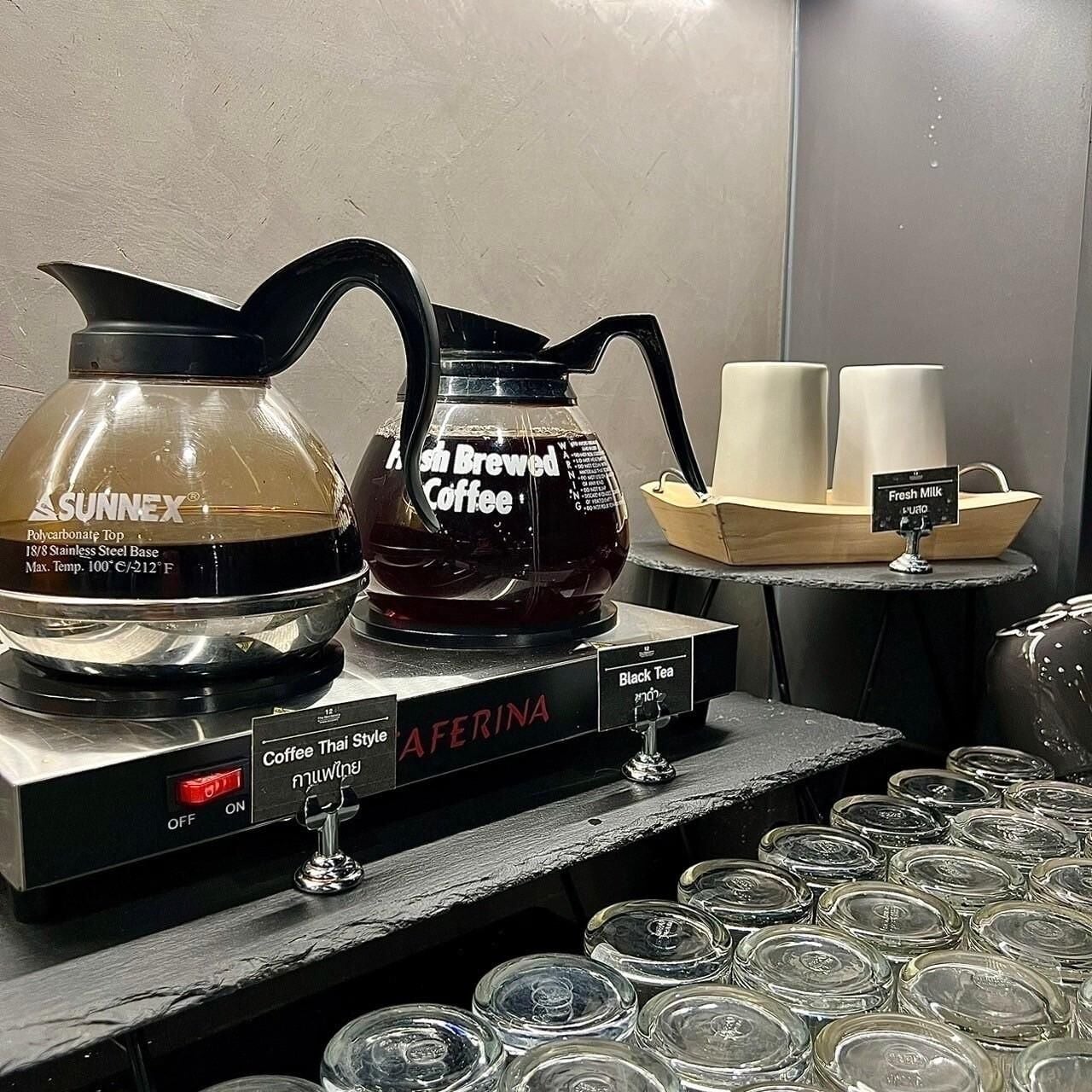 A coffee station with a SUNNEX coffee maker and a black tea dispenser, a fresh milk station, and a display of coffee cups.