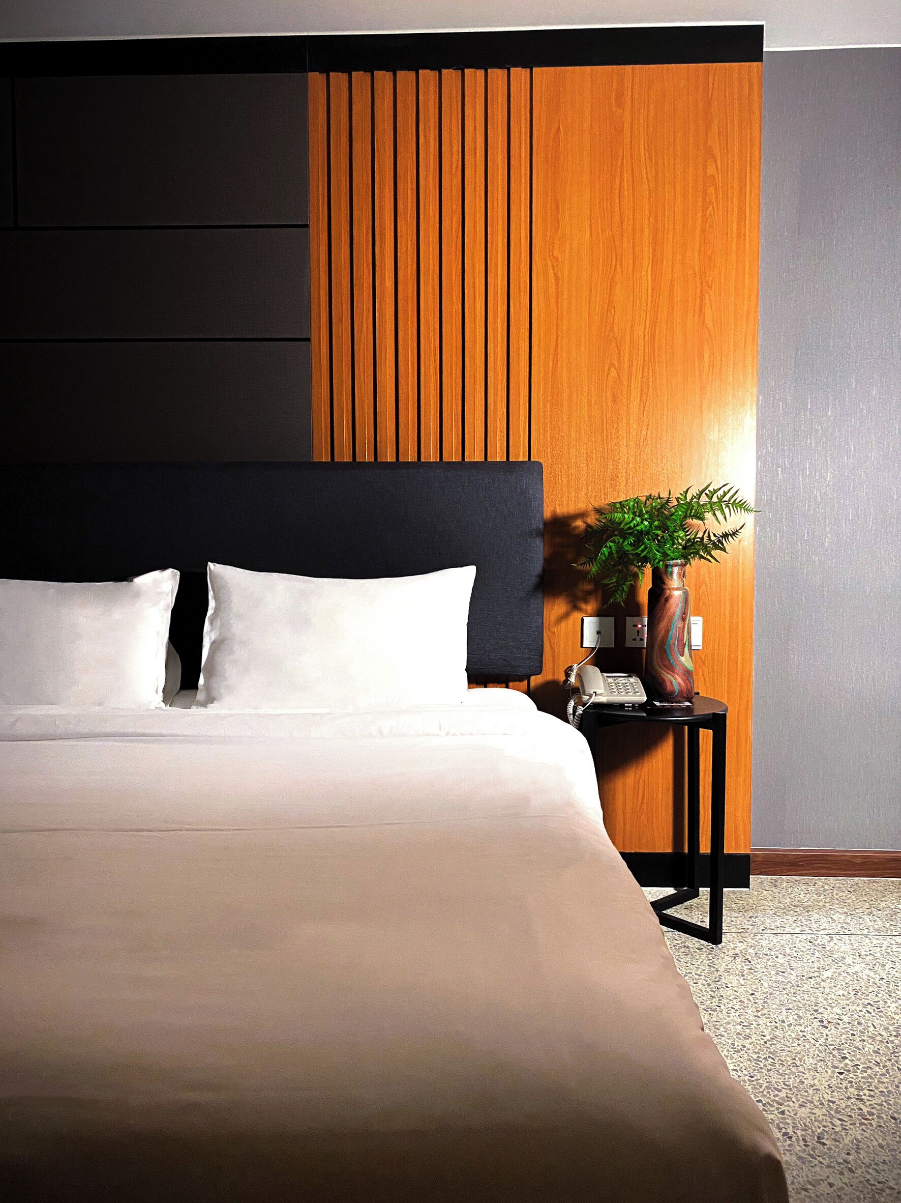 A modern hotel room with a large bed, dark headboard, wooden panel accent wall, and a small side table with a plant.