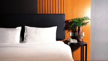 A modern hotel room with a large bed, dark headboard, wooden panel accent wall, and a small side table with a plant.