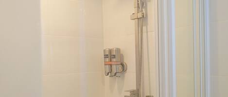 A modern shower with a handheld showerhead and a fixed showerhead, a glass door, and a small shelf with toiletries.