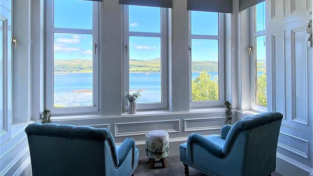 A room with three large windows offering a view of a body of water and greenery. There are two blue armchairs and a small round table with a vase.