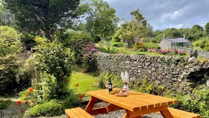 A wooden picnic table with benches in a garden setting, surrounded by lush greenery and a stone wall.