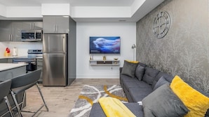 A modern kitchen with stainless steel appliances, a dining area with a bar stool, and a living room with a gray sofa and yellow cushions. - Elegant Apartment,  5mins from the Falls (Niagara Falls)