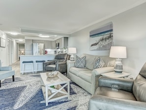 Un salon moderne avec un canapé gris, une table basse blanche et un tapis à motifs. On aperçoit une cuisine à l’arrière-plan, avec des meubles blancs et un coin repas.
