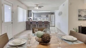 Un comedor con una mesa dispuesta para cuatro, una cocina al fondo y un ventilador de techo.