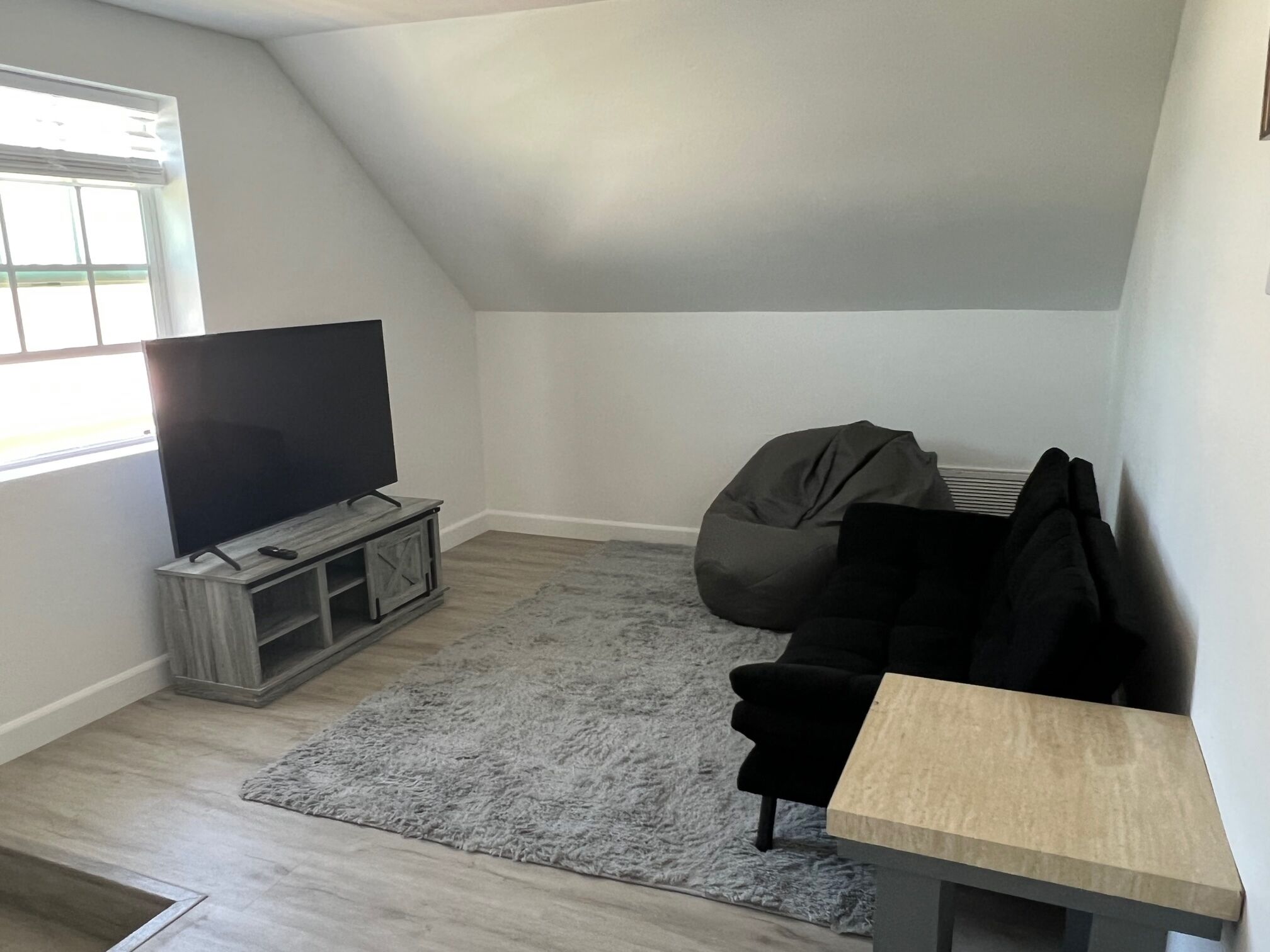 A modern living room with a flat-screen TV on a wooden stand, a grey shag rug, a black bean bag chair, and a small wooden table.