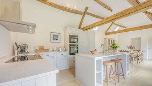 Een ruime keuken met witte kasten, een centraal eiland met krukken, houten balken in het plafond en een eethoek met stoelen.