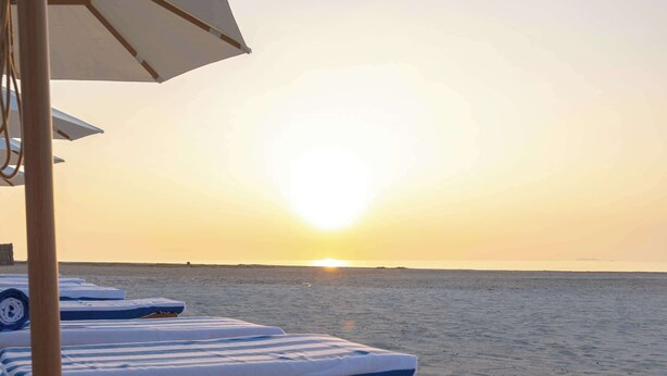 Een loungeruimte aan het strand met ligbedden en een opgerolde handdoek, met uitzicht op de zonsondergang boven de oceaan.