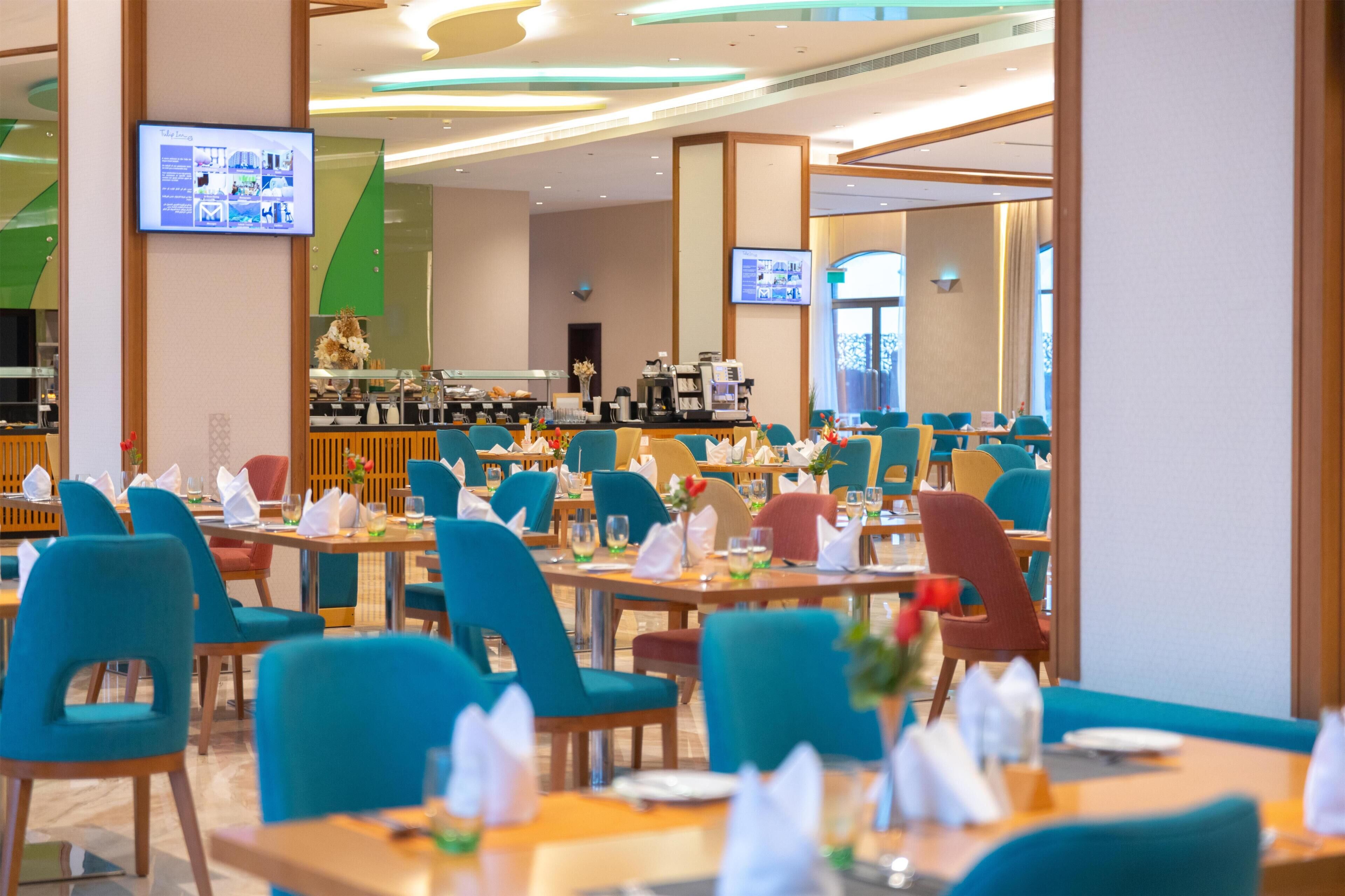 Un espace repas moderne doté de plusieurs tables et chaises, d’un coin café et d’écrans numériques fixés aux murs.