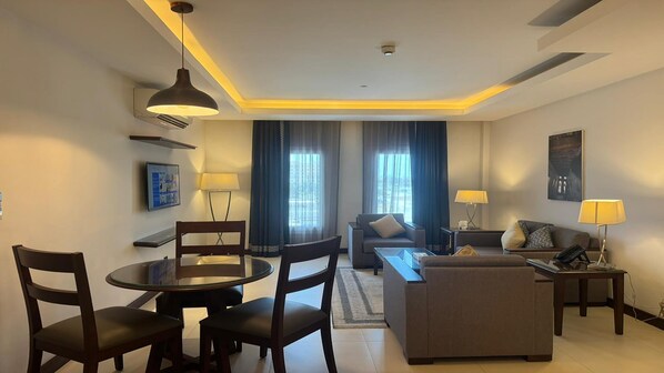 A modern hotel room with a dining area, a sofa, a coffee table, and a TV mounted on the wall.