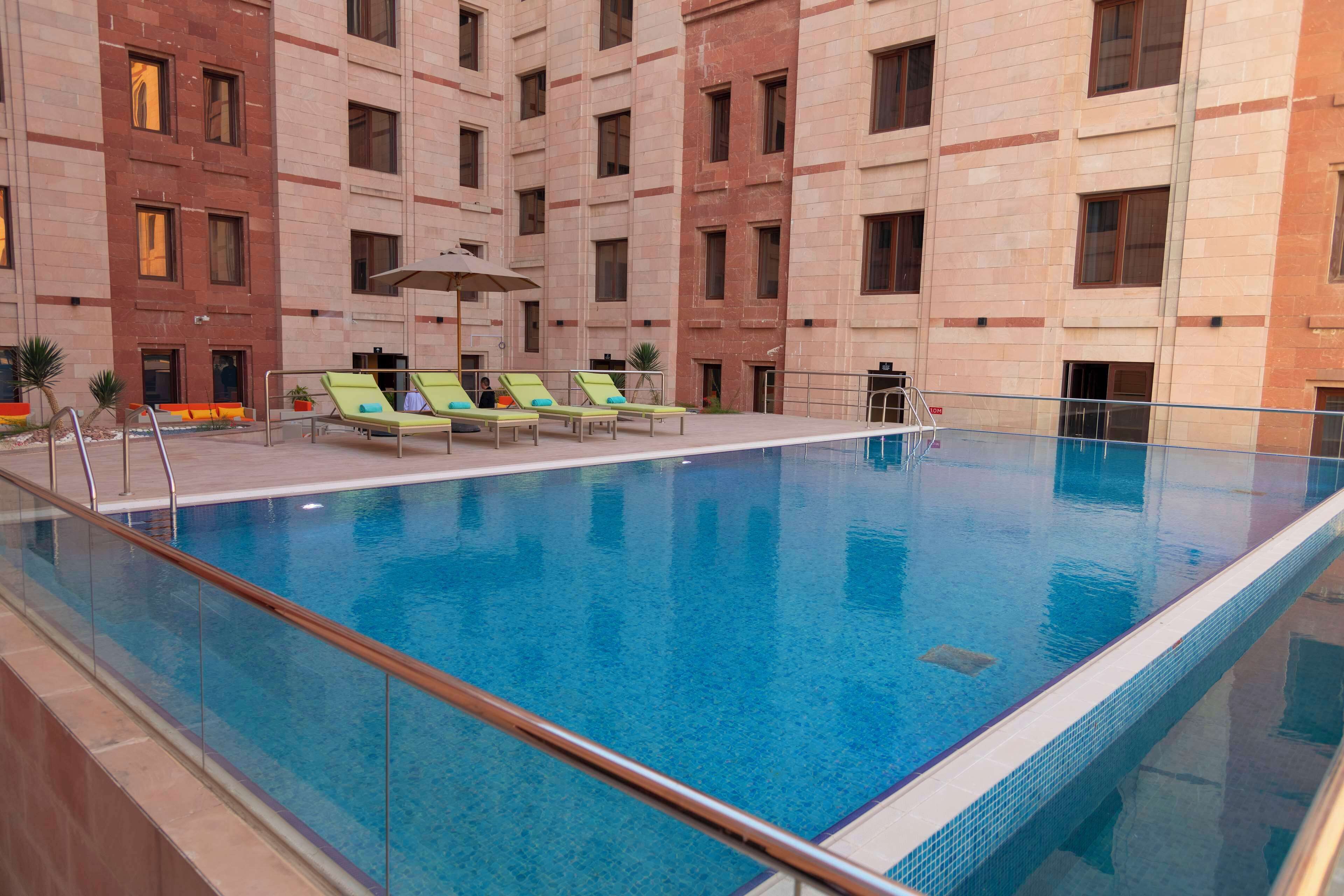 An indoor swimming pool with clear blue water, surrounded by a tiled pool deck, and a seating area with lounge chairs and a sun umbrella.