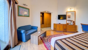 A hotel room with a blue sofa, a wooden desk, a bed with a striped bedspread, a television, and a wooden wardrobe. - Timber Trail Resort (Kasauli)