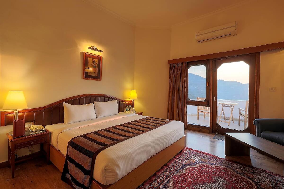 Une chambre d’hôtel avec un lit, des lampes de chevet, un bureau avec un téléphone, une chaise et une vue à travers une porte vitrée.