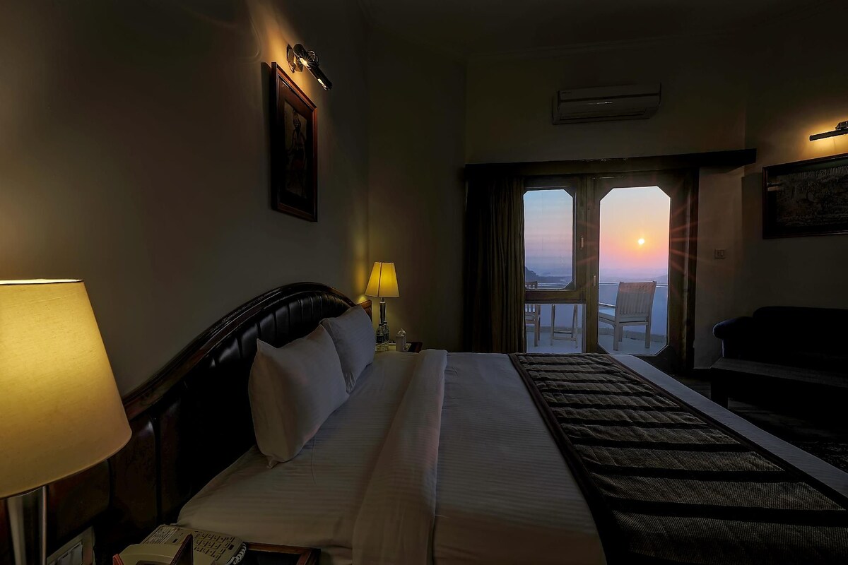 A hotel room with a bed, two bedside lamps, a desk with a telephone, and a view of the sunset through a door.