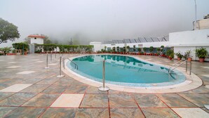 A swimming pool with a tiled deck, surrounded by potted plants and a small building with a red roof. - Timber Trail Resort (Kasauli)