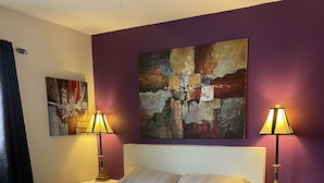 Ein ordentlich bezogenes Bett mit weißem Kopfteil, zwei Nachttische mit Lampen und ein großes abstraktes Gemälde an der Wand.