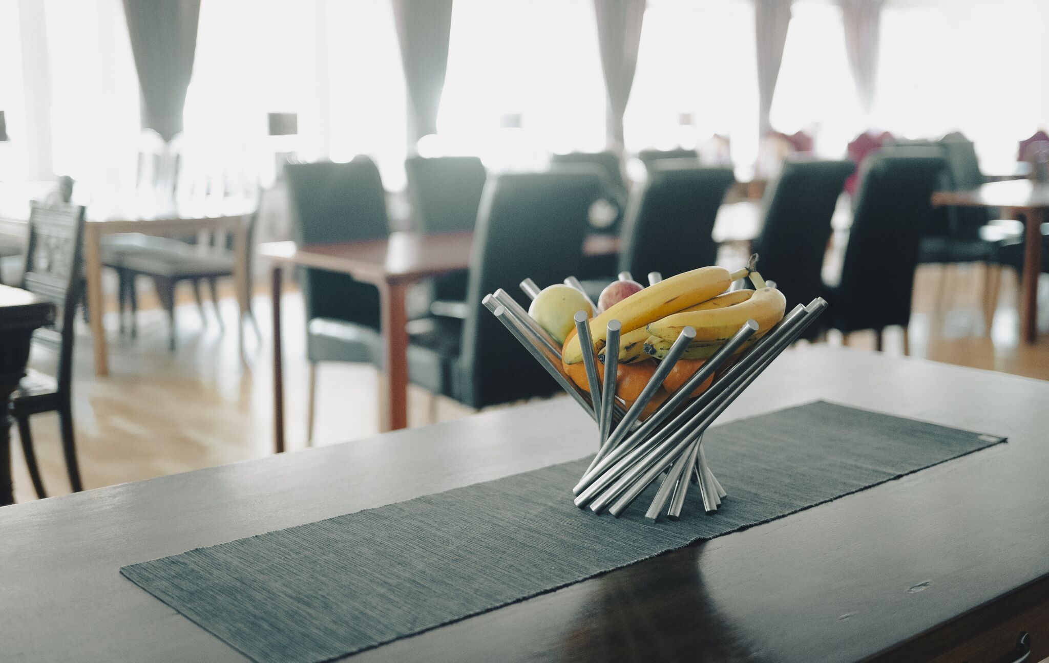 A dining table with a metal fruit holder containing bananas and apples, set on a dark placemat in a well-lit room with chairs and tables in the background.