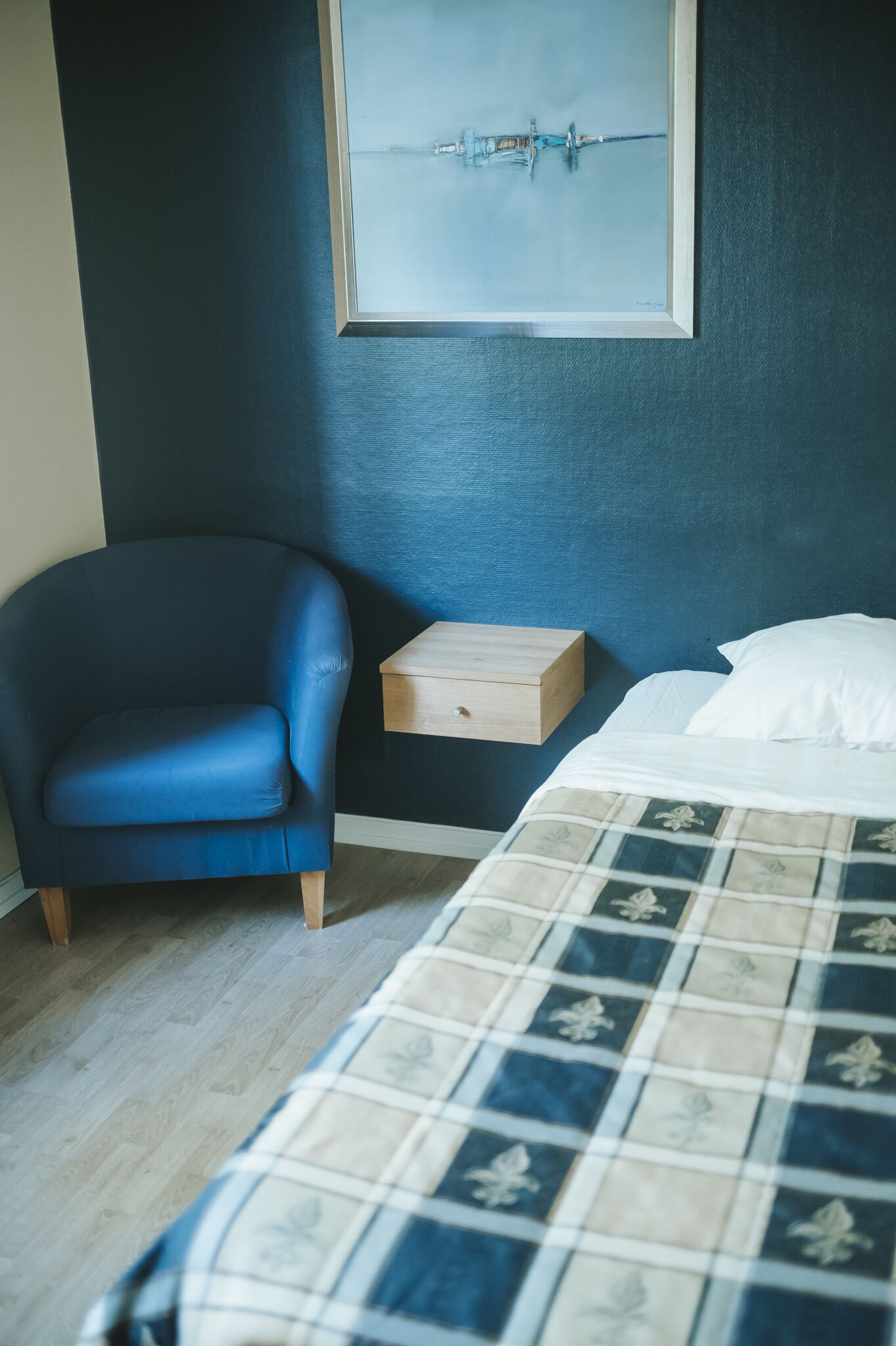 Une chambre à coucher avec un lit, un fauteuil bleu, une table de chevet en bois et un tableau accroché au mur.