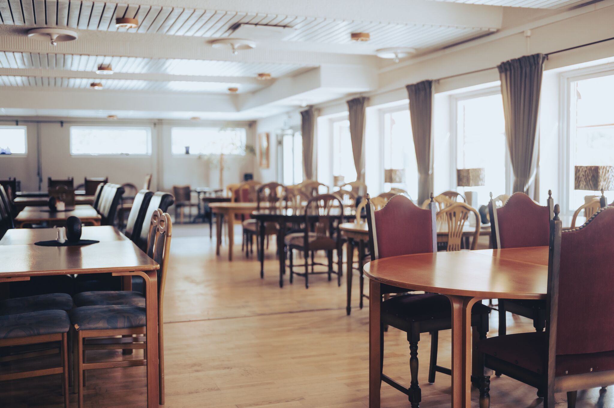 A spacious dining area with multiple wooden tables and chairs, large windows with curtains, and a high ceiling with exposed beams.