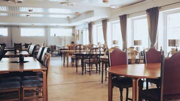 A spacious dining area with multiple wooden tables and chairs, large windows with curtains, and a high ceiling with exposed beams.
