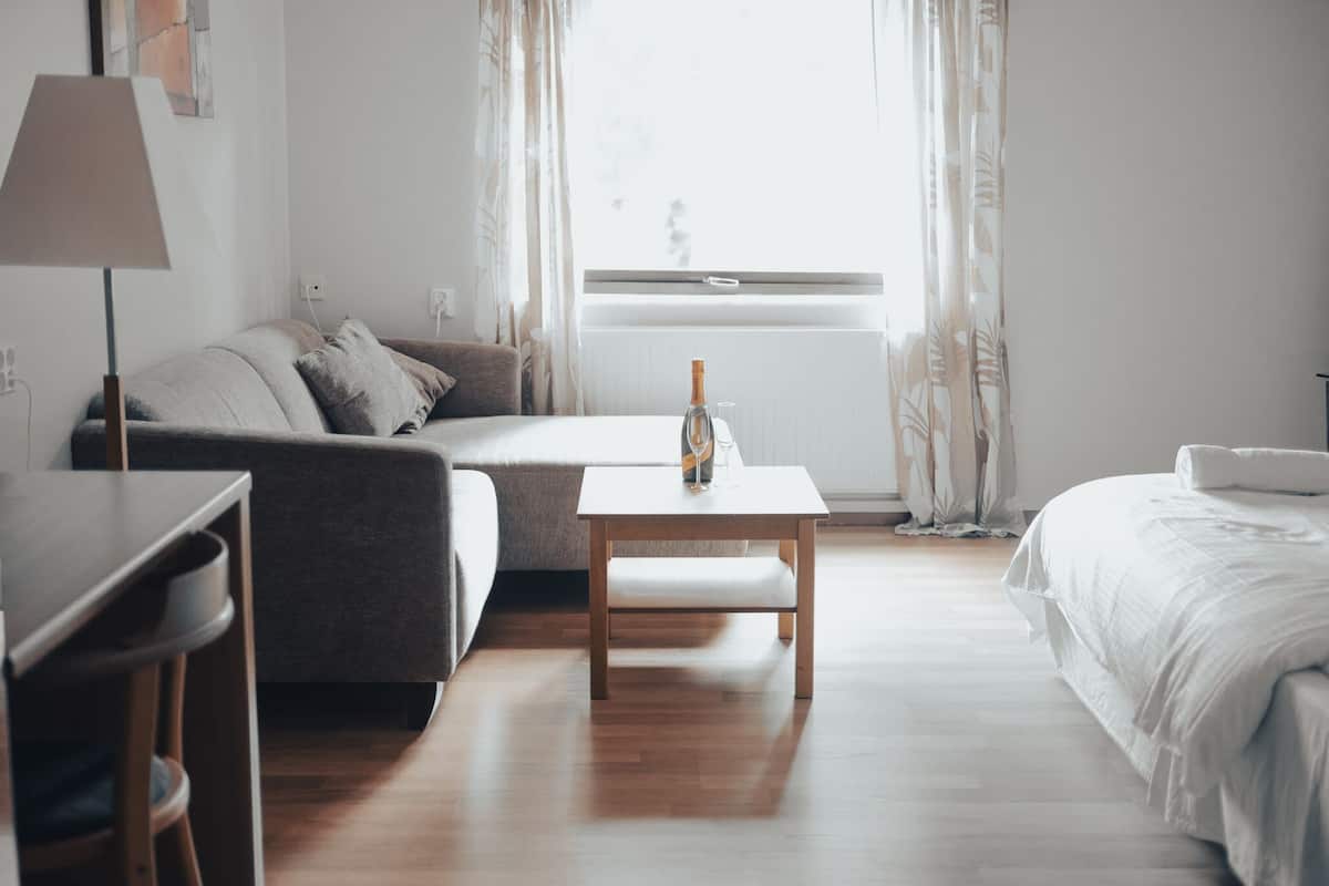 A modern hotel room with a wooden floor, a sofa, a small table with a bottle, a bed, and a lamp.