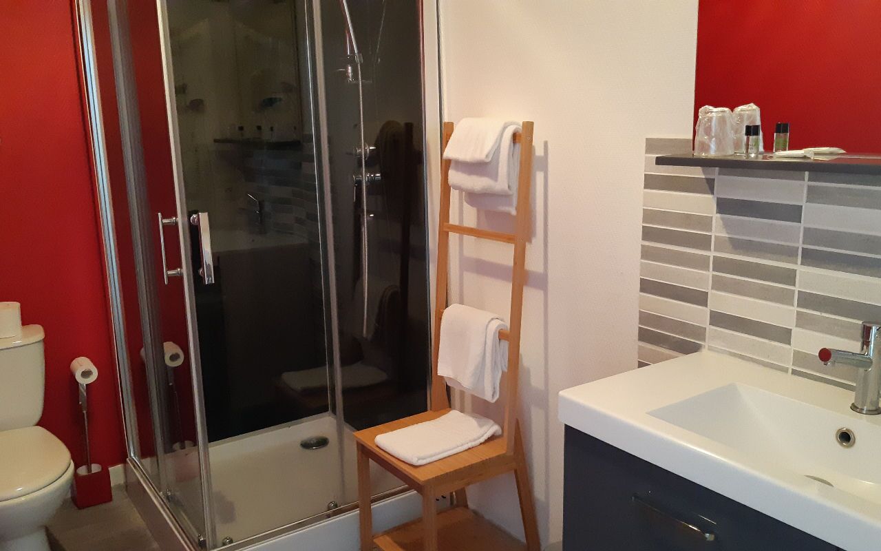 Une salle de bain avec une douche à parois de verre, une chaise en bois avec une serviette, un lavabo blanc et un mur d’accent rouge.