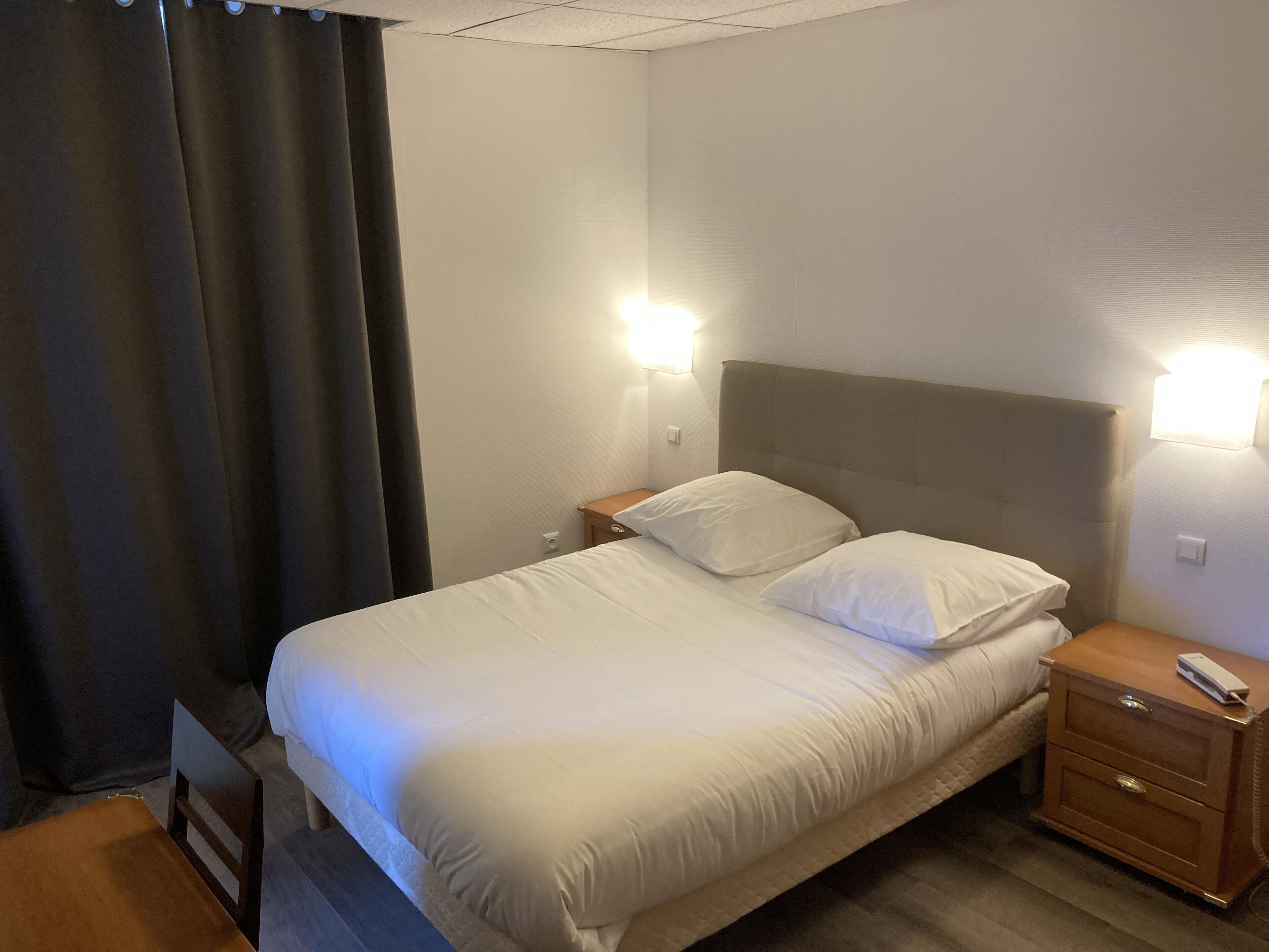 A neatly made bed with white linens, a wooden nightstand with a remote control, and two wall-mounted lights.