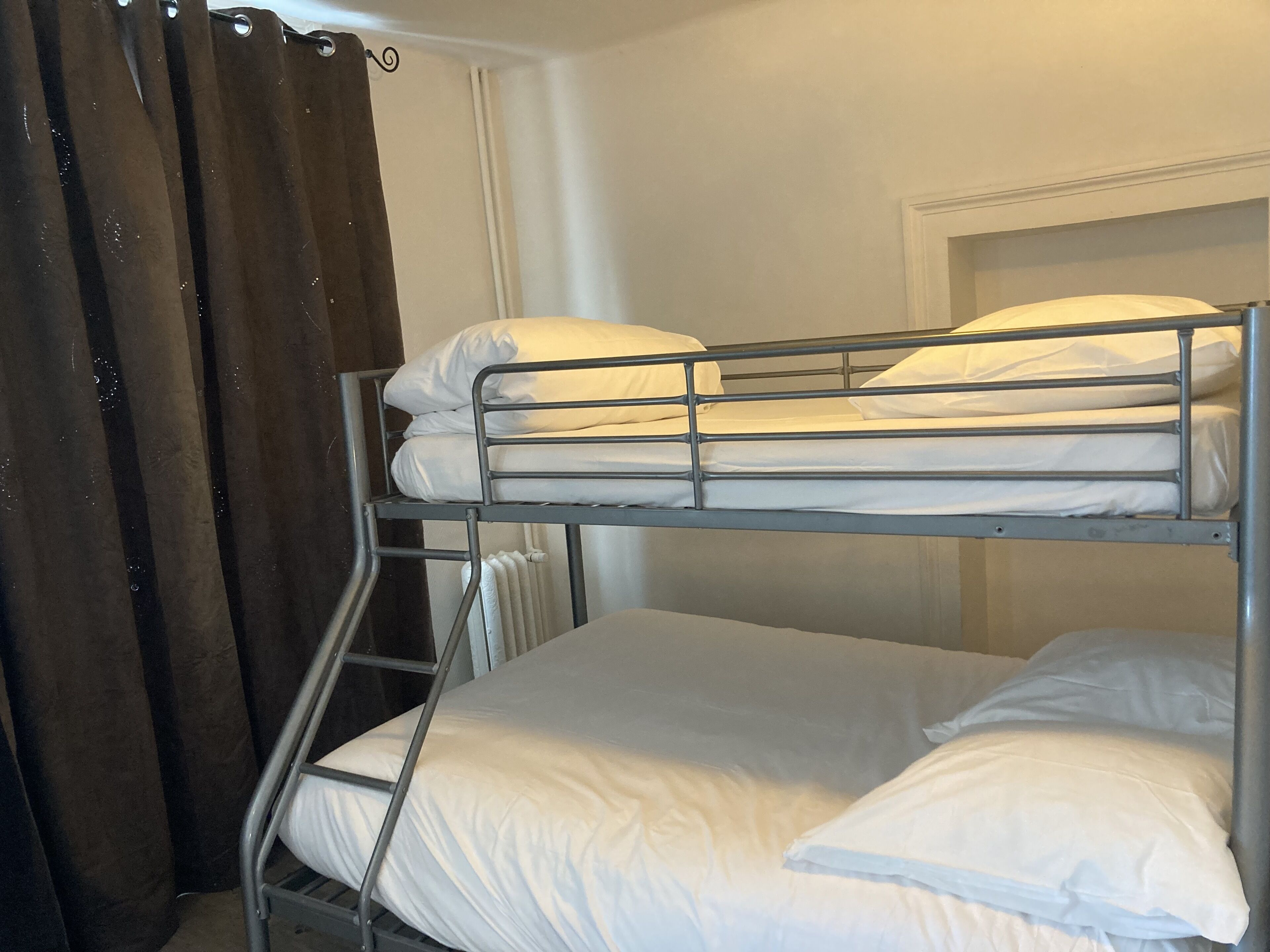 Un lit superposé avec une literie blanche, dans une pièce dotée d’un rideau et d’un radiateur.