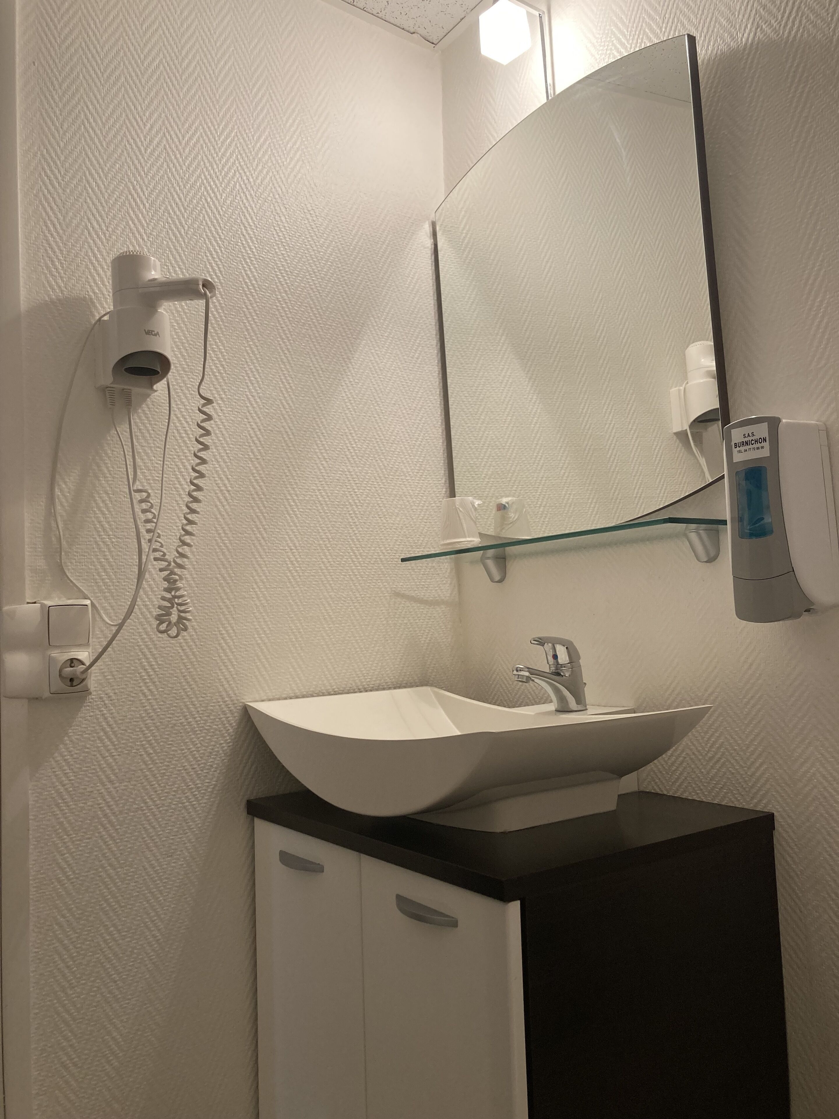 A bathroom with a white sink, a mirror, a hairdryer, and a glass shelf.