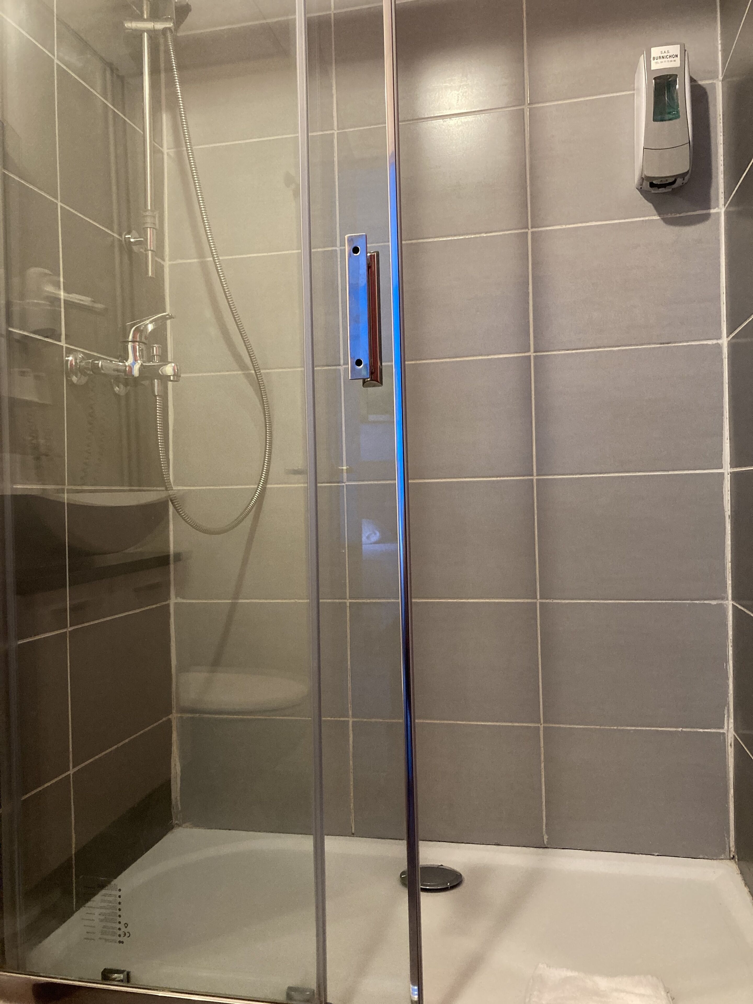 A glass-enclosed shower stall with a handheld showerhead and a wall-mounted hand dryer.