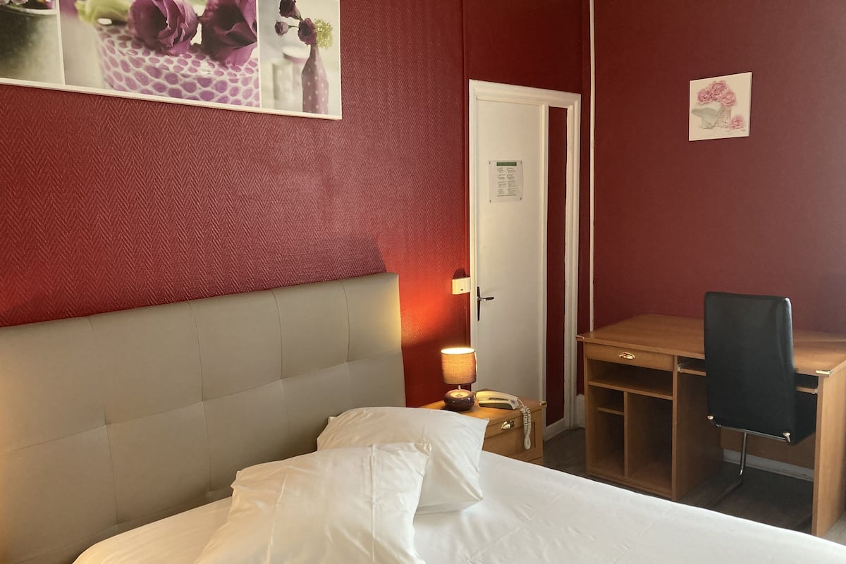 Ein Hotelzimmer mit Bett, Schreibtisch, Stuhl, Nachttisch mit Lampe und einer Tür.