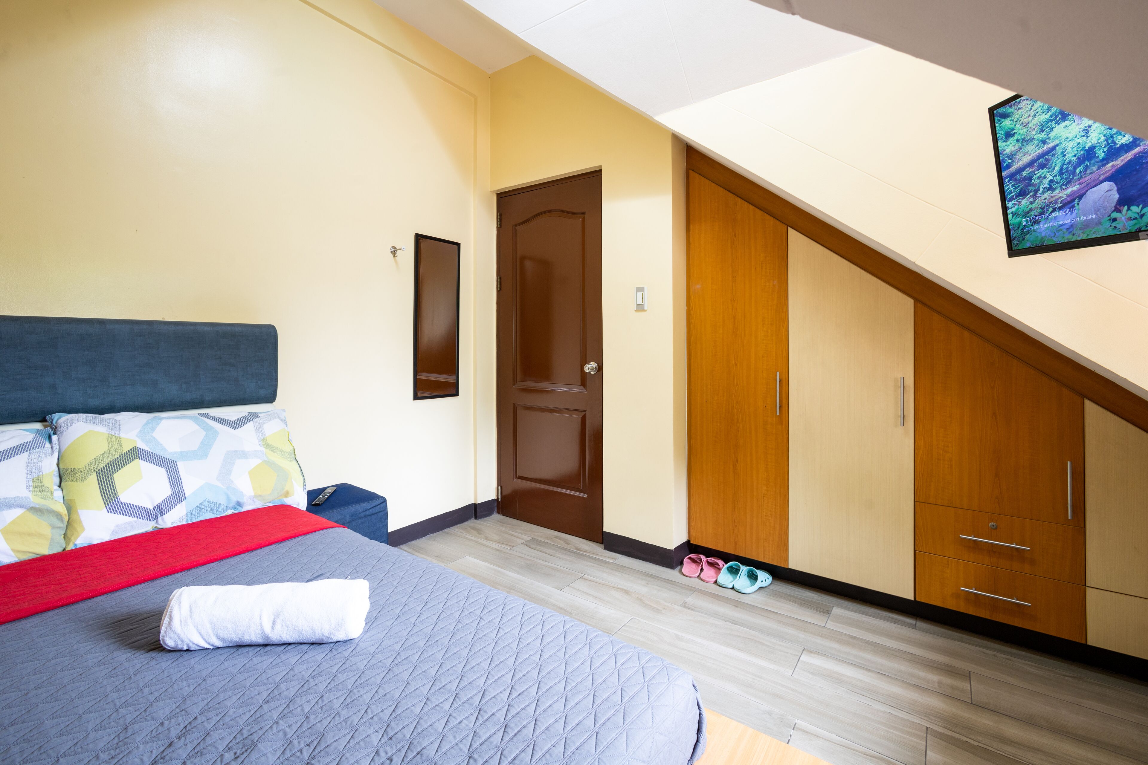 A bedroom with a bed, a red throw blanket, a neatly folded towel, a wooden wardrobe, and a pair of slippers.