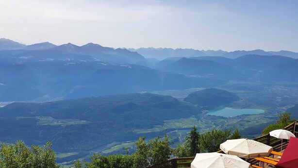 A mountain view with a lake, a wooden fence, and outdoor seating with umbrellas. - PanoramaHotel Penegal (Ruffre)