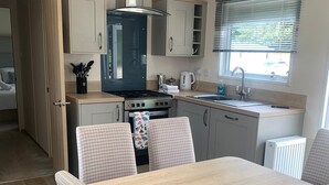 A compact kitchen with a stove, sink, and cabinets, a dining table with chairs, and a window with blinds. - Feorag Lodge -  Retreat Group (Aviemore)