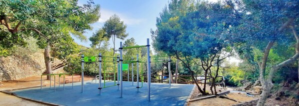 An outdoor gym area with exercise equipment, surrounded by trees and a stone wall. - Villa Rakhi by Solidays - Moraira, Costa Blanca (Moraira)