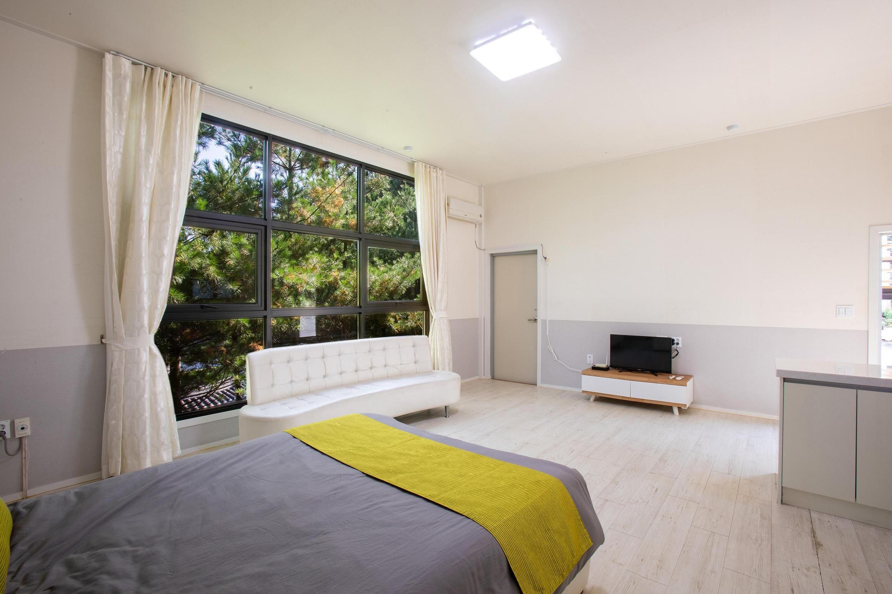 A modern bedroom with a large window, a bed with a grey and yellow bedspread, a white sofa, a TV on a wooden stand, and a small kitchenette area.
