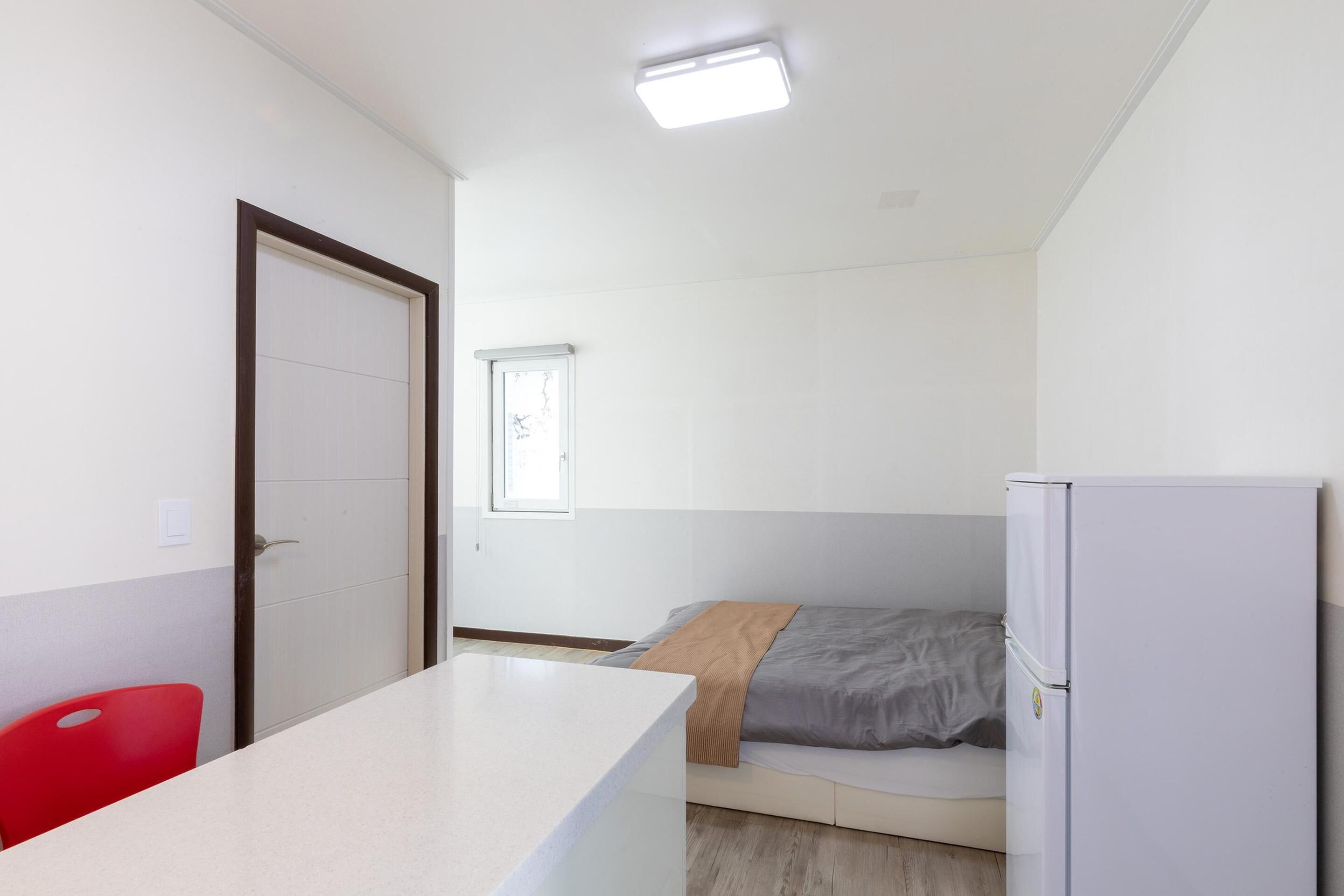 Ein kleiner, sauberer Raum mit einem Bett, einem weißen Kühlschrank, einem roten Stuhl und einem Fenster mit einer Karte an der Wand.