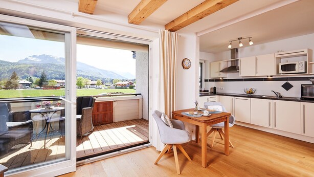 A modern kitchen with white cabinets, a wooden dining table, and a view of a mountainous landscape through large glass doors. - Ferienwohnung Alpenzauber mit Balkon, ca. 60 qm (Reit im Winkl)