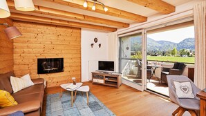A cozy living room with a fireplace, a TV, and a large sliding glass door leading to an outdoor area with a view of mountains. - Ferienwohnung Alpenzauber mit Balkon, ca. 60 qm (Reit im Winkl)