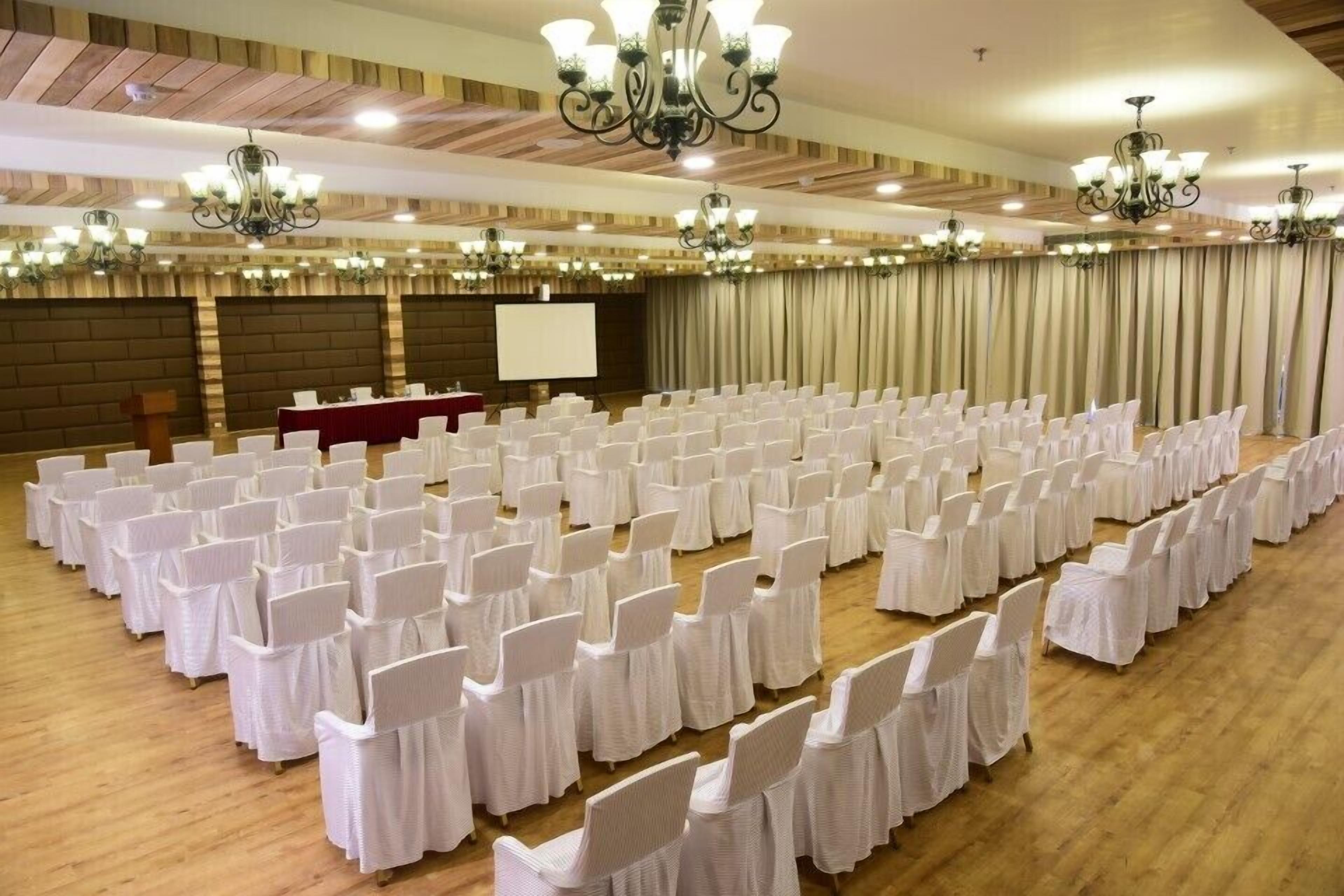 Un salón de conferencias con filas de sillas blancas, una pantalla de proyector y una mesa roja.