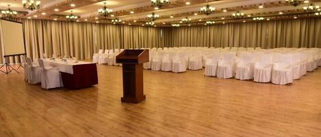 Un amplio salón de conferencias con filas de sillas blancas, un atril y una pantalla a la izquierda.