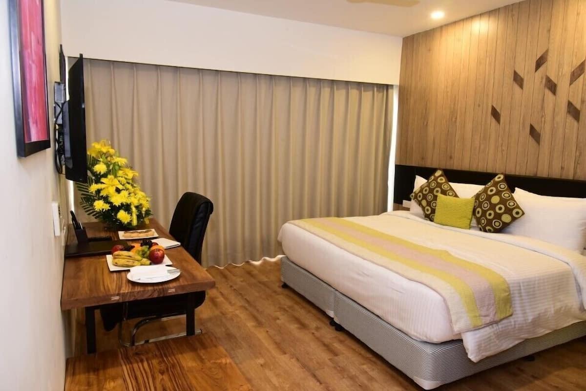 Ein Hotelzimmer mit einem Bett, einem Schreibtisch mit einem Laptop, einem Sessel und einem Esstisch mit Früchten und einer Blumenvase.