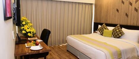 A hotel room with a bed, a desk with a laptop, a chair, and a dining table with fruits and a vase of flowers.