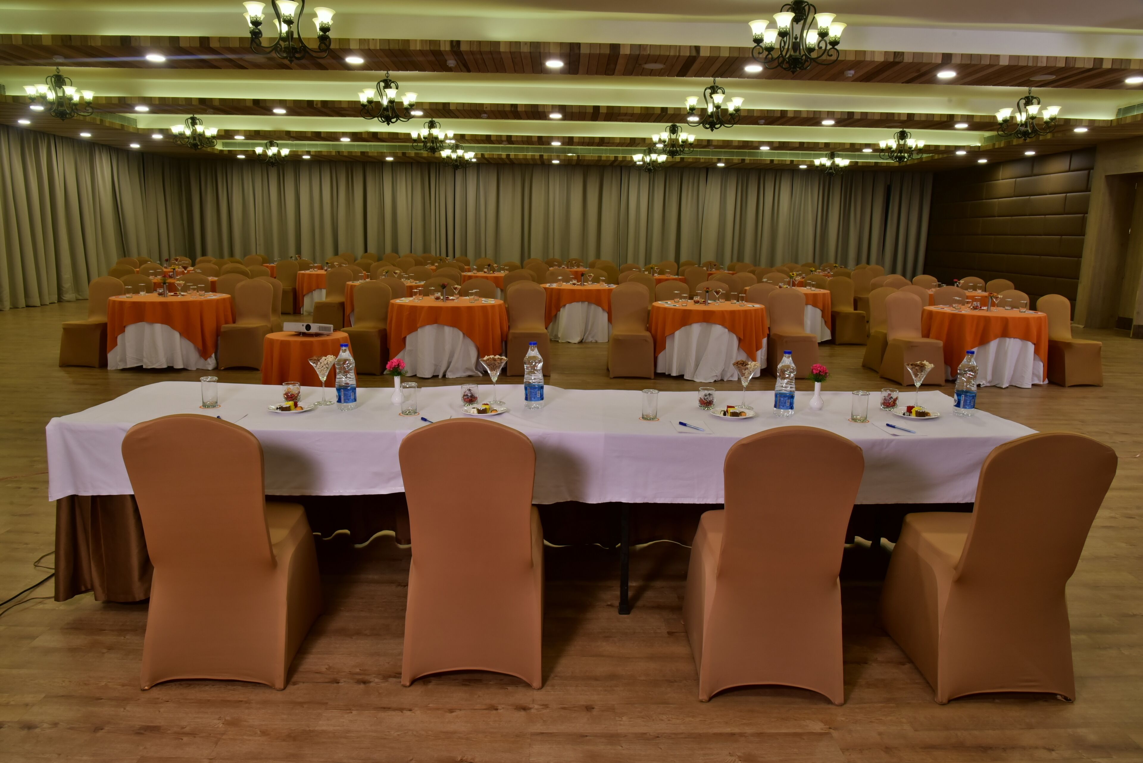 A banquet hall with round tables covered in white tablecloths, orange chairs, and chandeliers on the ceiling.
