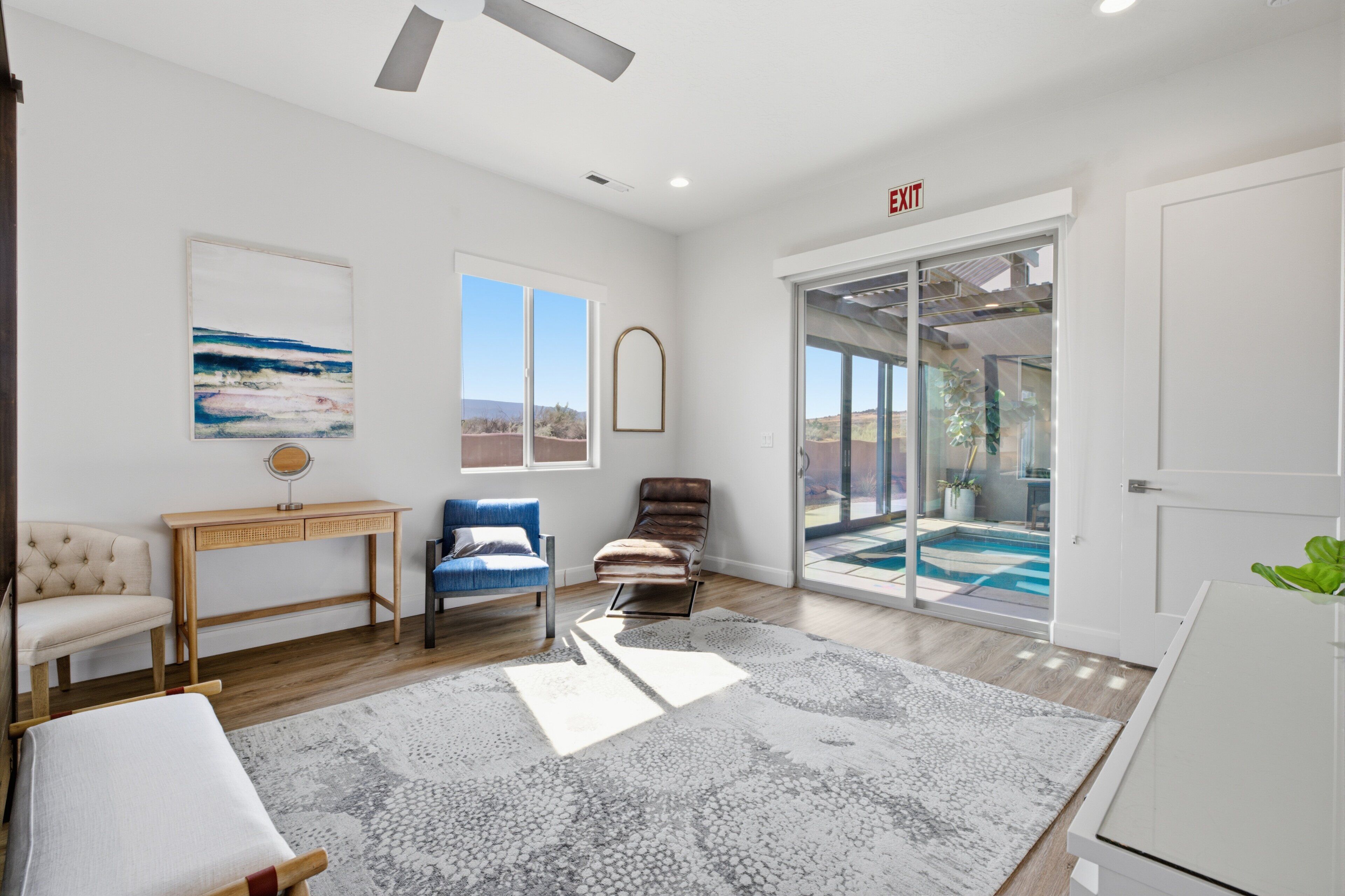A spacious living room with a large window, a sliding glass door leading to a pool, a wooden desk, and a comfortable seating area.