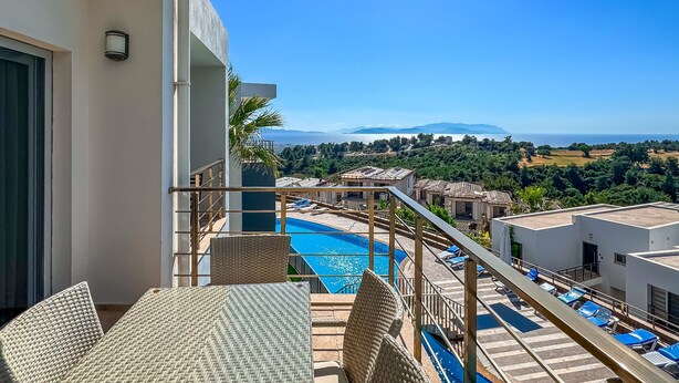 A balcony with wicker furniture overlooking a swimming pool and a scenic view of the sea and mountains.