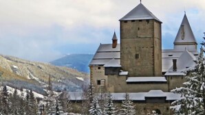 A historic building with a central tower and two smaller towers, surrounded by snow-covered trees and a mountainous backdrop.