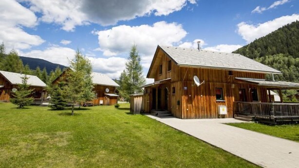 A wooden cabin with a large porch, surrounded by a grassy area and trees, with mountains in the background.