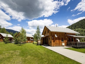 Une cabane en bois dotée d’une grande véranda, entourée d’une pelouse et d’arbres, avec des montagnes en arrière-plan.