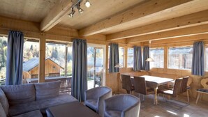 A cozy wooden cabin with a sofa, chairs, and a dining table. Large windows with curtains offer views of the surrounding area.